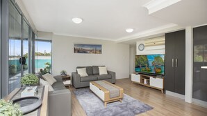 A modern living room with a grey sofa, a coffee table, a flat-screen TV, and a large window overlooking a pool. - Spacious. Coastal. Fresh. (Forster)