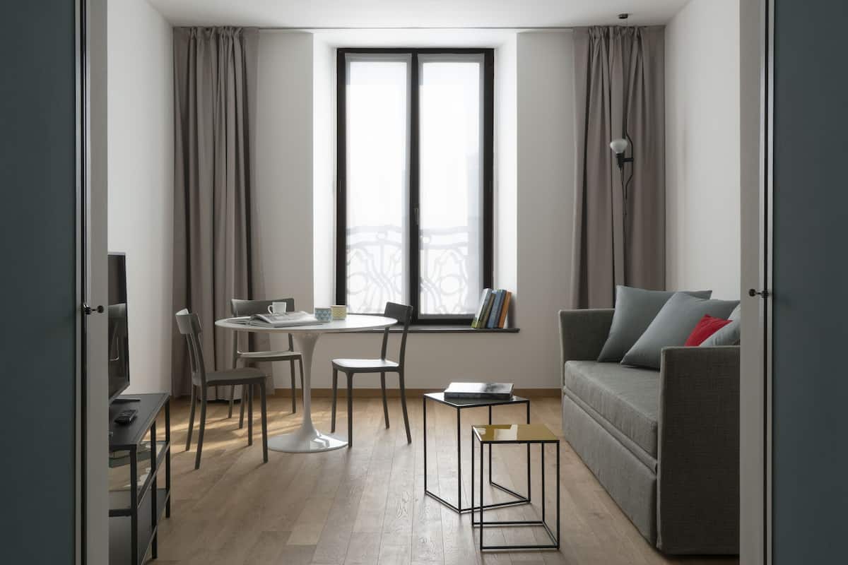 A modern living room with a grey sofa, a small coffee table, a dining table with chairs, and a window with curtains.