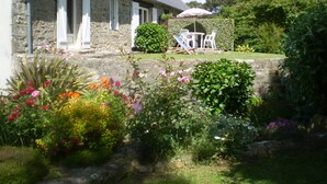 Un edificio de piedra con techo inclinado, un pequeño patio amoblado al aire libre y un jardín con diversas plantas y flores.