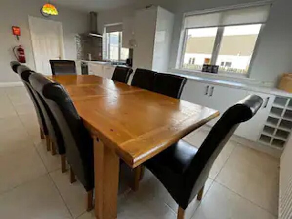 A dining area with a wooden table and black chairs, a kitchen in the background, and a window with a view.