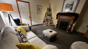 Ein Wohnzimmer mit einer Couch, einem Hocker und einem Sessel, einem Weihnachtsbaum, einem Kamin und einem gerahmten Bild an der Wand.