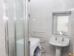A bathroom with a glass shower enclosure, a washing machine, a sink with a mirror, and a window with a view.