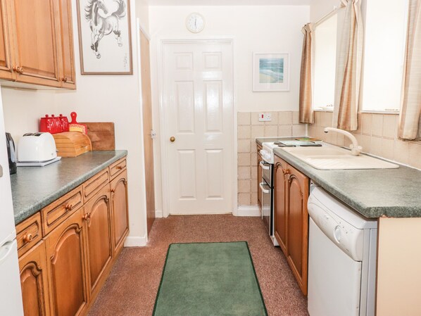 A compact kitchen with wooden cabinets, a green rug, a white door, and a small window with curtains. - Gwylan Apartment (Tenby)