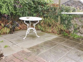 Mesa metálica redonda con tapa blanca y patas ornamentadas, ubicada en un patio de piedra rodeado de exuberante follaje y un muro de piedra.
