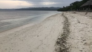 En strand med en gångbana av betong, en byggnad med tak av halm och berg i fjärran.