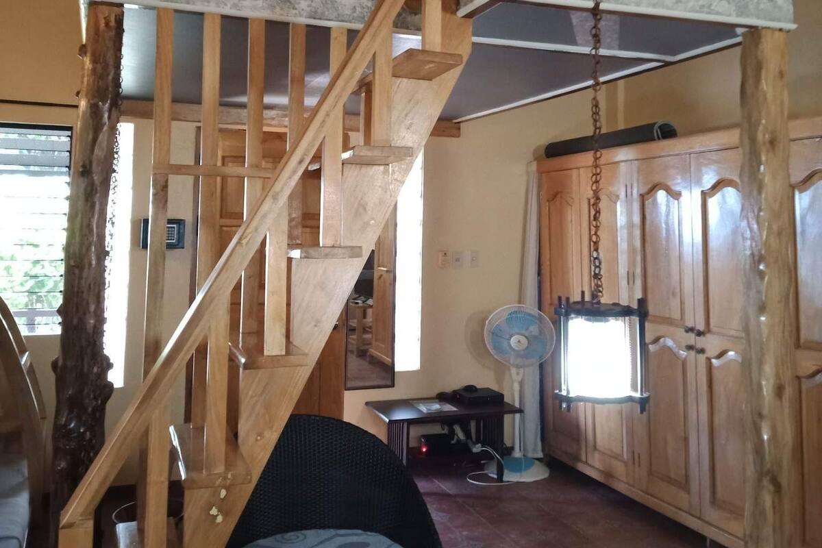 Una escalera de madera de diseño rústico, un ventilador y una silla de mimbre en una habitación con paredes de madera y suelo de baldosas.