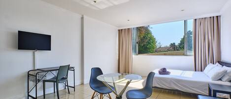 Una habitación de hotel moderna con cama, televisión, escritorio y sillas, y ventana con cortinas.
