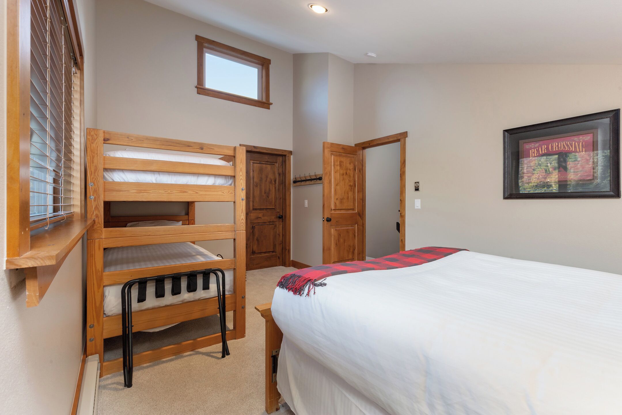 A bedroom with a bunk bed, a single bed, a window with blinds, a door, and a framed picture on the wall.