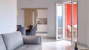 A modern living room with a grey sofa, a dining area with a table and chairs, a kitchen with white cabinets, and a sliding glass door leading to an outdoor area with red walls. - Apartment Flores by Interhome (Rabac)