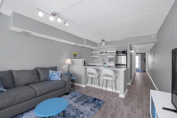 A modern living room with a grey sofa, a blue coffee table, and a kitchen area with white bar stools.