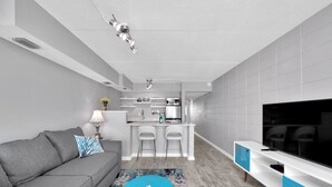 Een moderne woonkamer met een grijze bank, een blauwe salontafel, een televisie en een keuken met barstoelen.