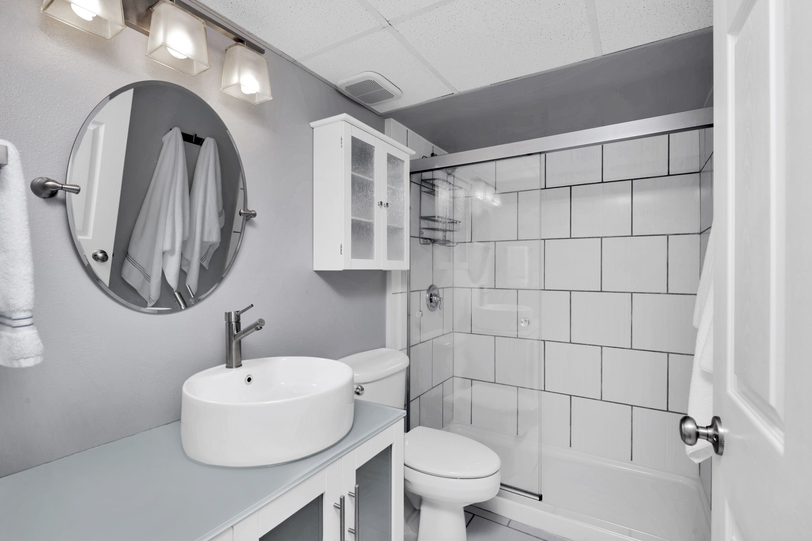Et moderne badeværelse med et rundt spejl, en hvid vask, et toilet og et bruseområde med glasvægge.