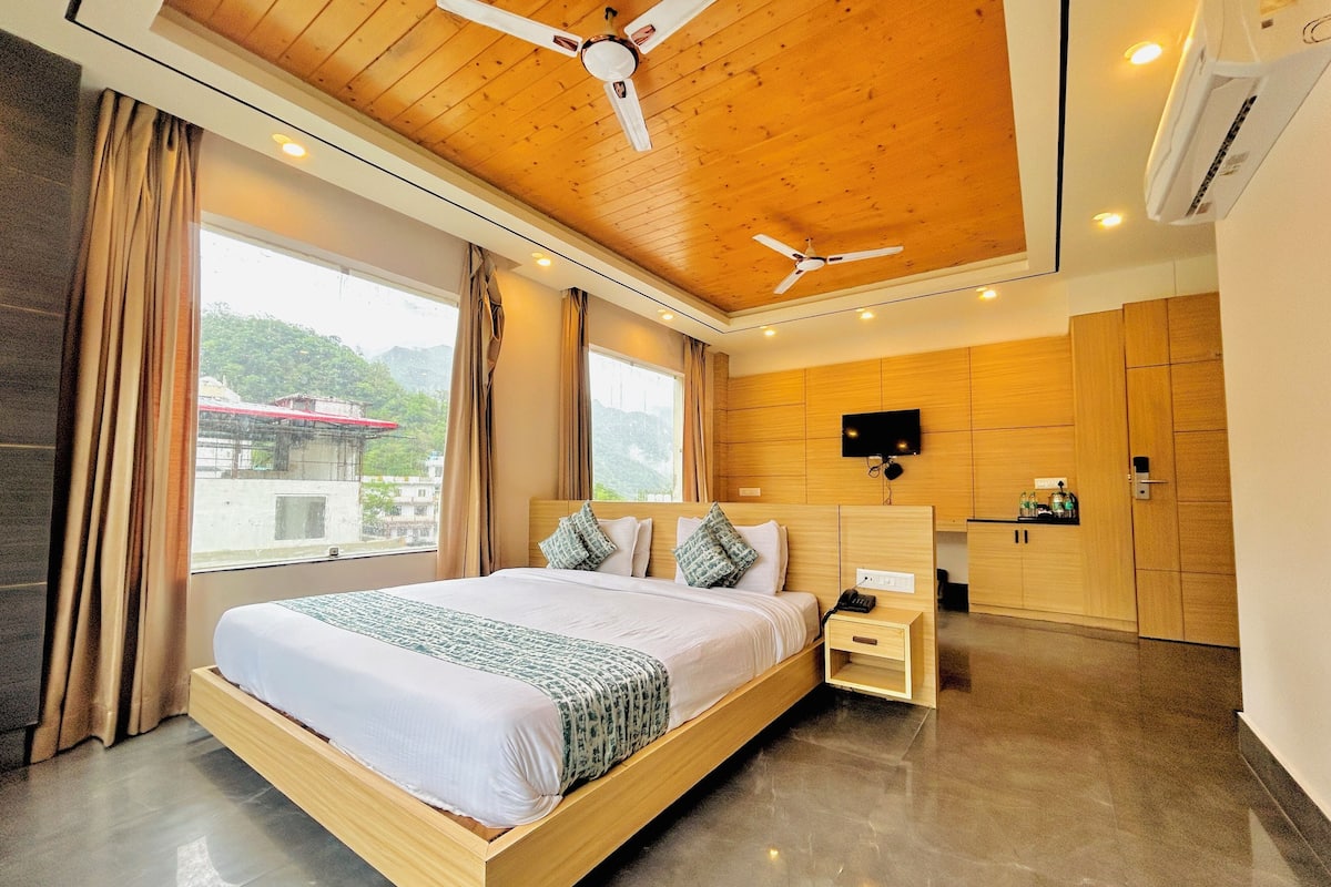 Una habitación de hotel moderna con techo de madera, una cama grande con ropa blanca, una mesita de noche, un televisor montado en la pared y vistas al exterior.