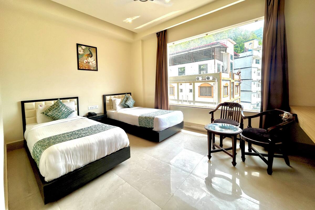 Habitación de hotel con dos camas, una silla, una mesa, un ventilador de techo y un ventanal con vistas a edificios.