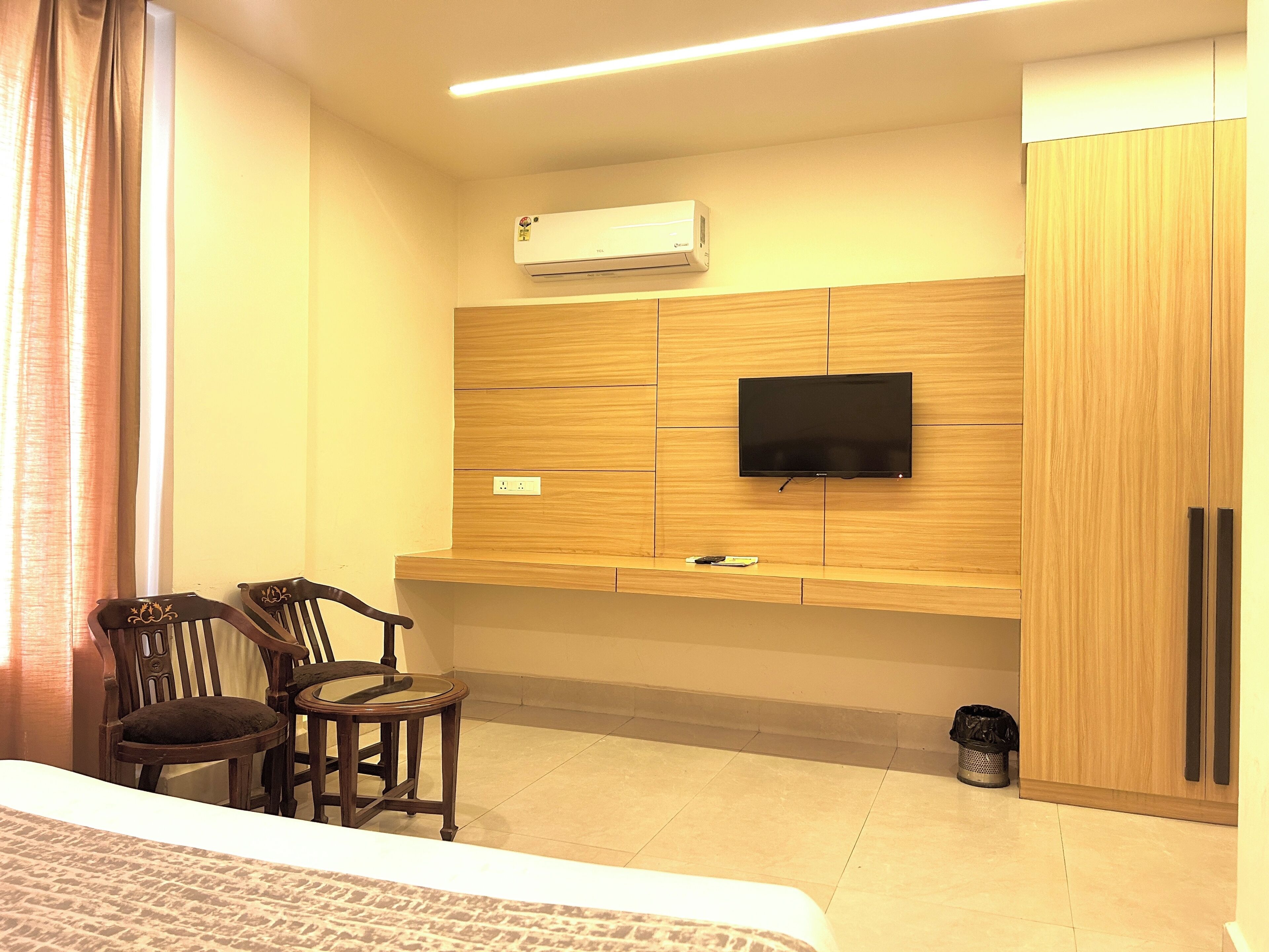 A hotel room with a bed, a desk with two chairs, a flat-screen TV mounted on a wooden panel, an air conditioner on the wall, and a trash bin.
