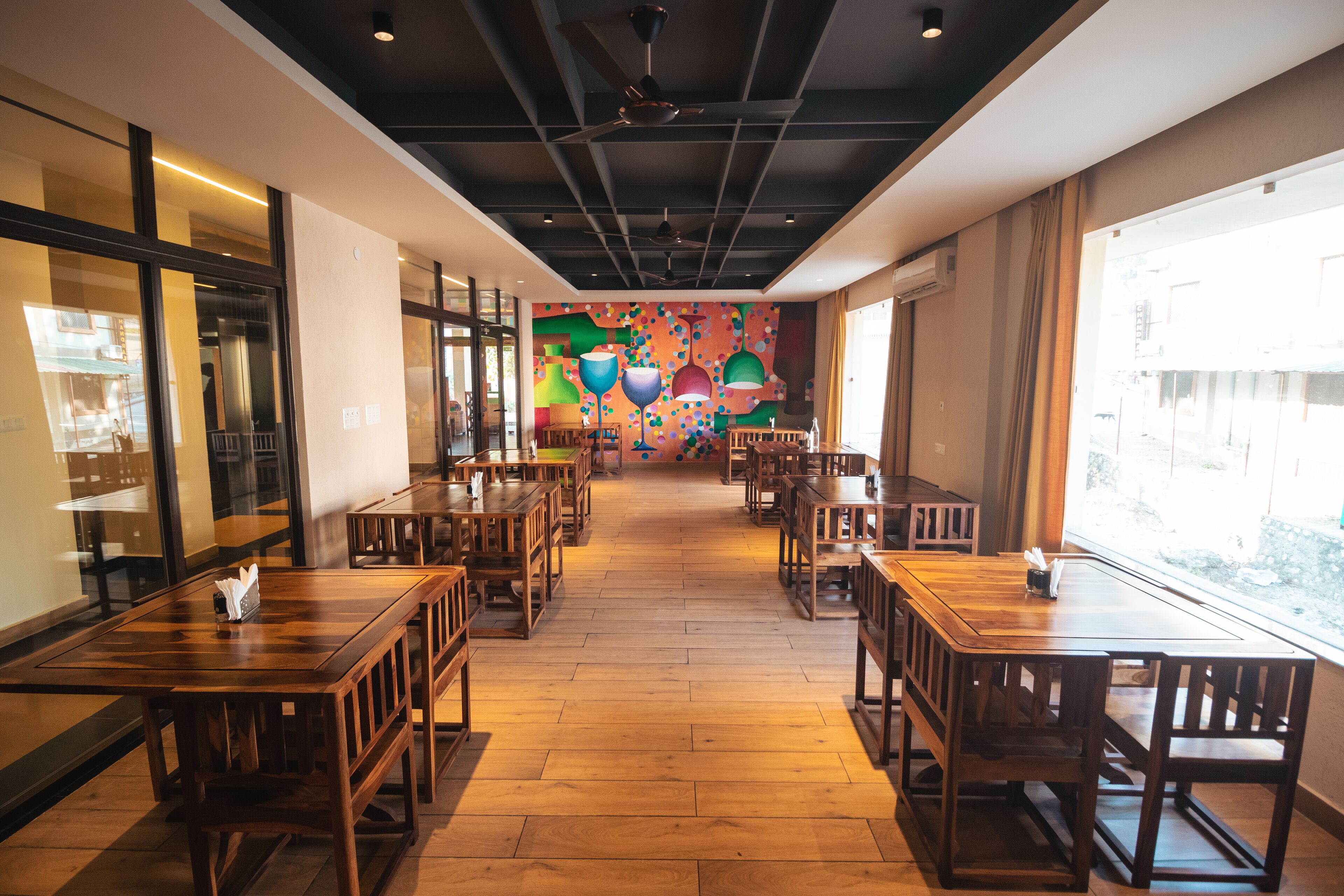 A restaurant with wooden tables and chairs, a colorful mural on the wall, and large windows allowing natural light.