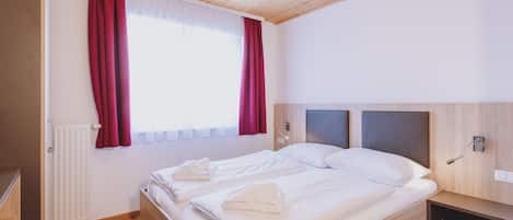 Un dormitorio con techo de madera, una cama con ropa blanca, una mesita de noche y una ventana con cortinas rojas.