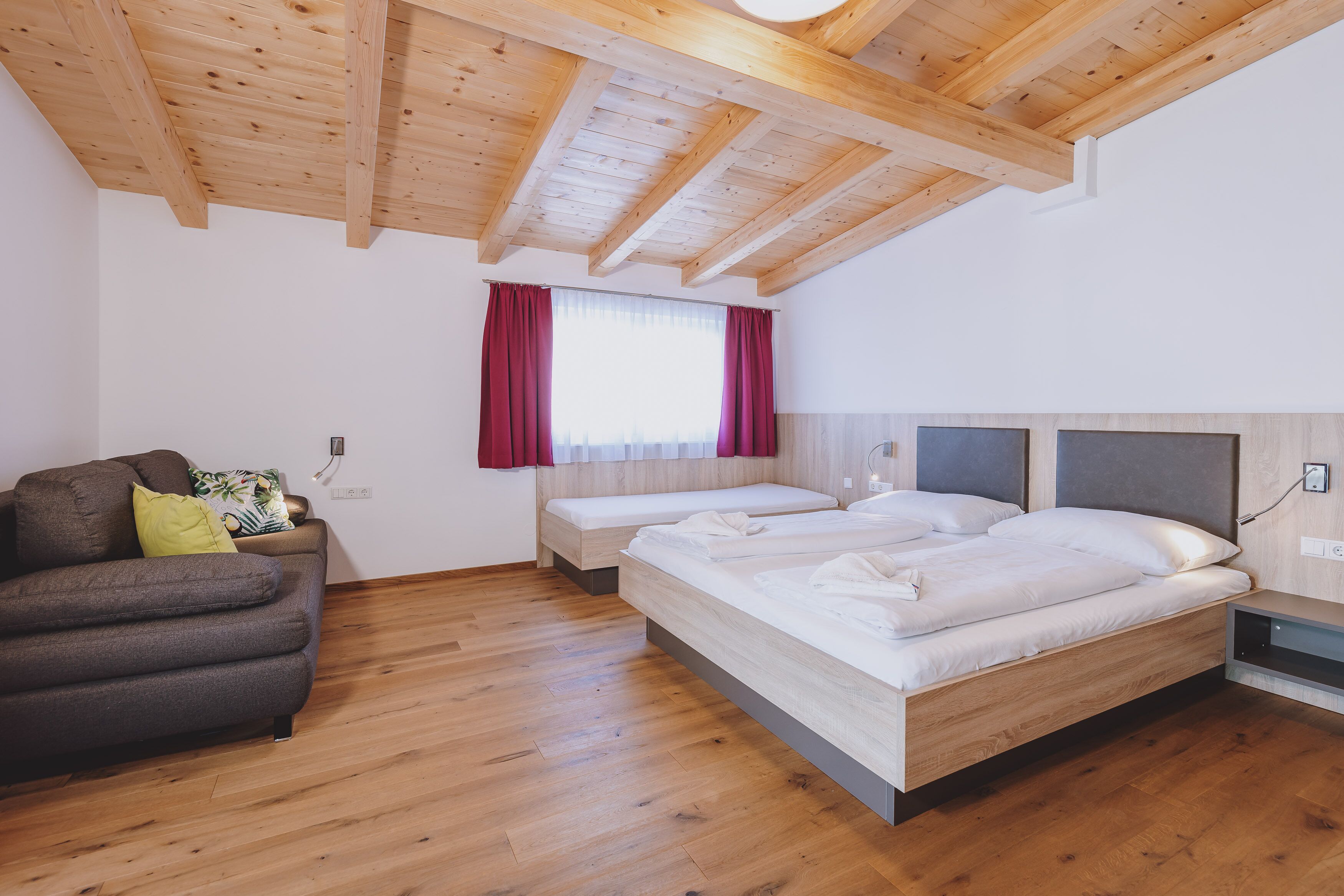 A room with wooden flooring, two beds with white linens, a sofa with cushions, and a window with red curtains.