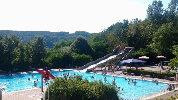 Una piscina con tobogán, rodeada de árboles y un paisaje montañoso.