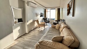 Una sala de estar moderna con un sofá color beige, un televisor y una ventana con vistas a un balcón.