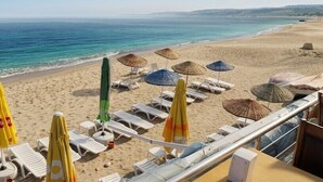 Un hotel frente a la playa con tumbonas y sombrillas, vista despejada al mar y una playa de arena.