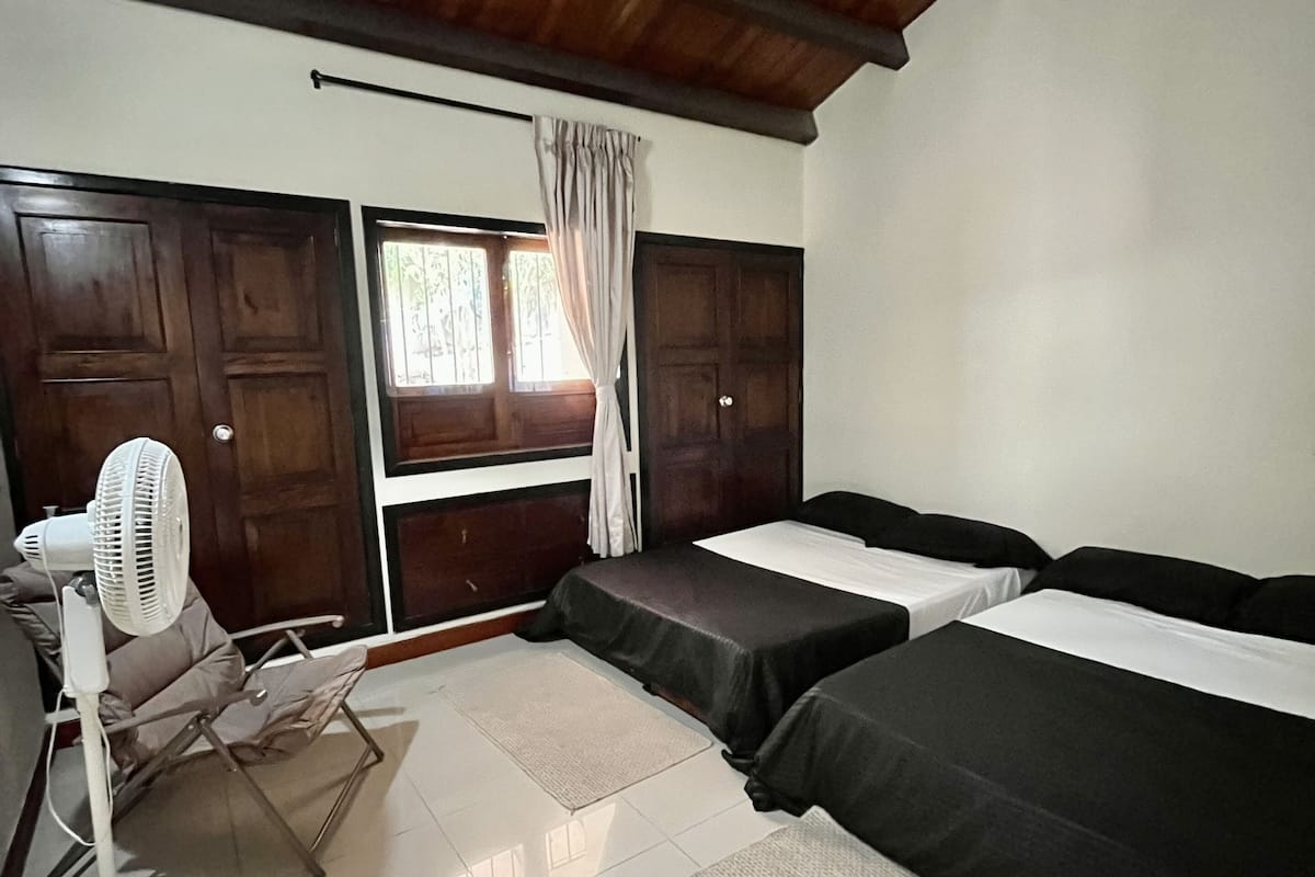 A room with two beds, a fan, a window with curtains, and a wooden ceiling.