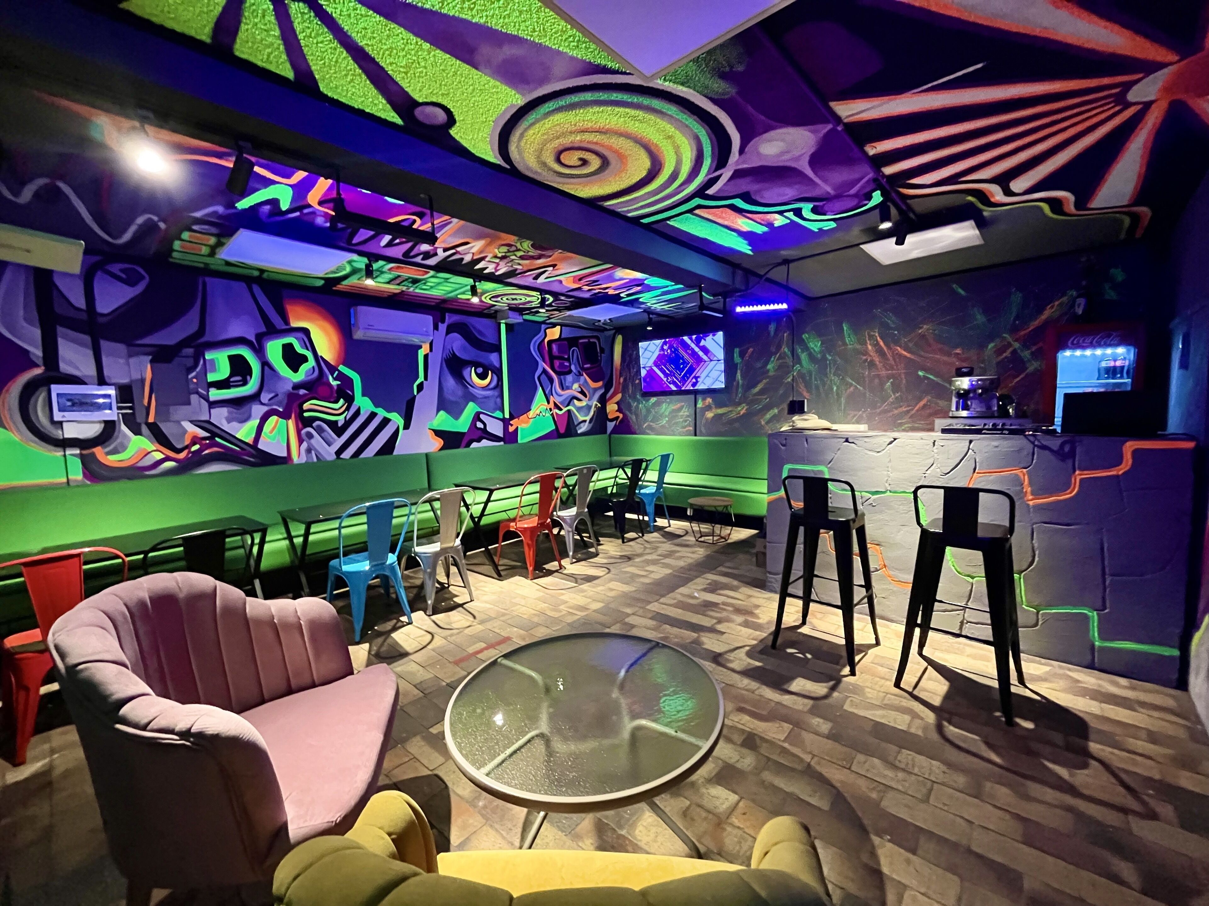 A vibrant bar area with colorful graffiti, a seating area with various chairs, and a bar counter with a Coca-Cola refrigerator.