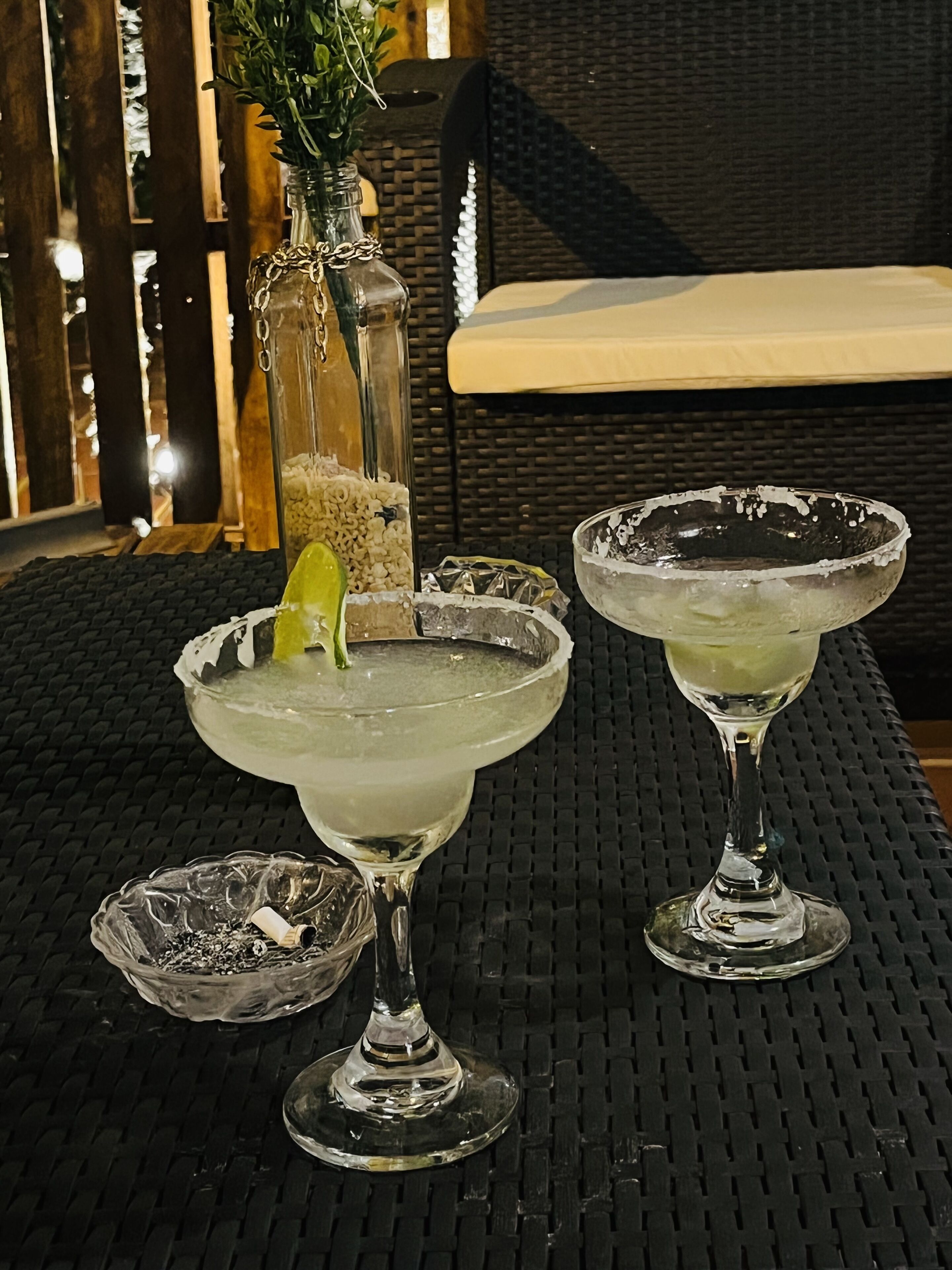 Two margaritas on a woven table with a wicker chair and a glass bottle in the background.
