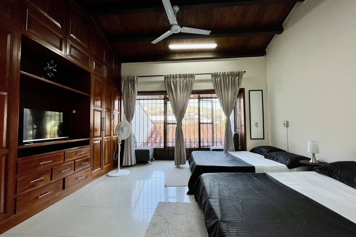 A hotel room with a large bed, a TV, wooden cabinets, a ceiling fan, and a view of the outside.