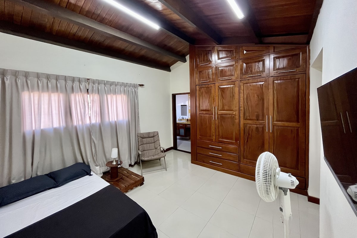 A bedroom with a bed, a chair, a desk, a wardrobe, and a fan.