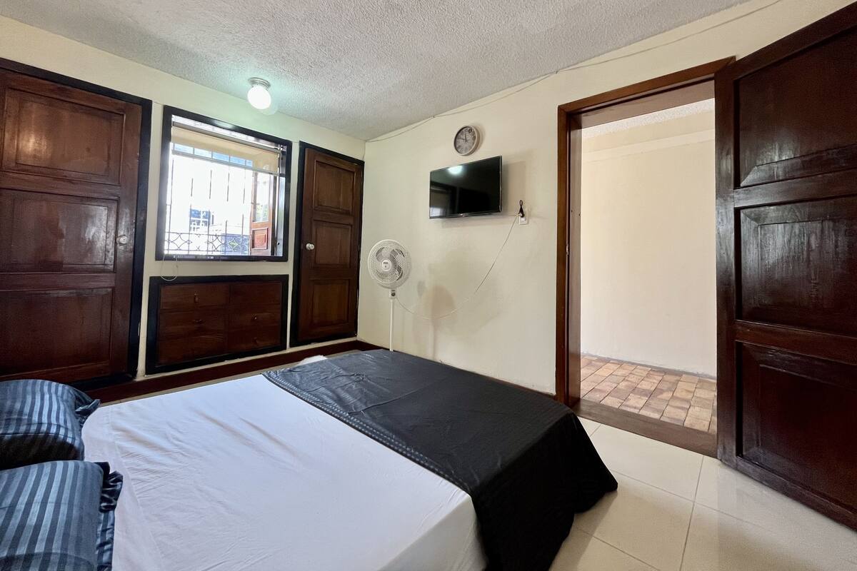 A bedroom with a bed, a TV, a fan, and a window.