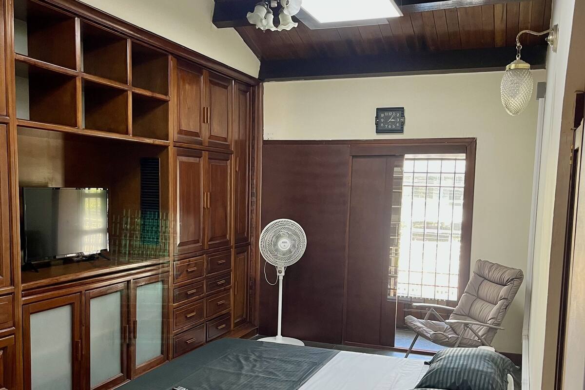 A bedroom with a bed, wooden wardrobe, ceiling fan, and a chair.