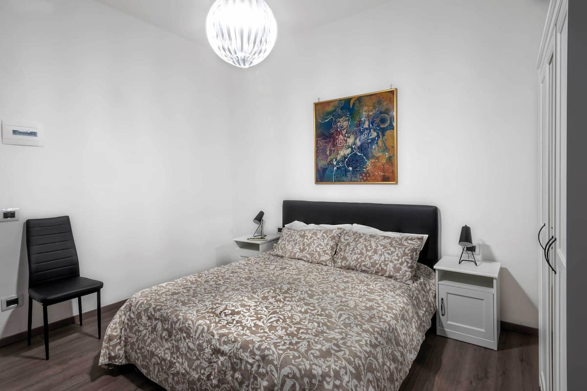 Un dormitorio con cama, una silla, una mesita de noche, una lámpara y un cuadro en la pared.