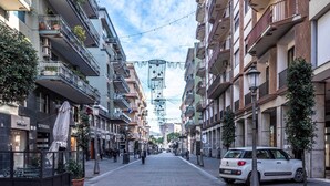 Una estrecha calle urbana bordeada de edificios de varios pisos, con balcones con plantas, un coche blanco estacionado y una acera para peatones.