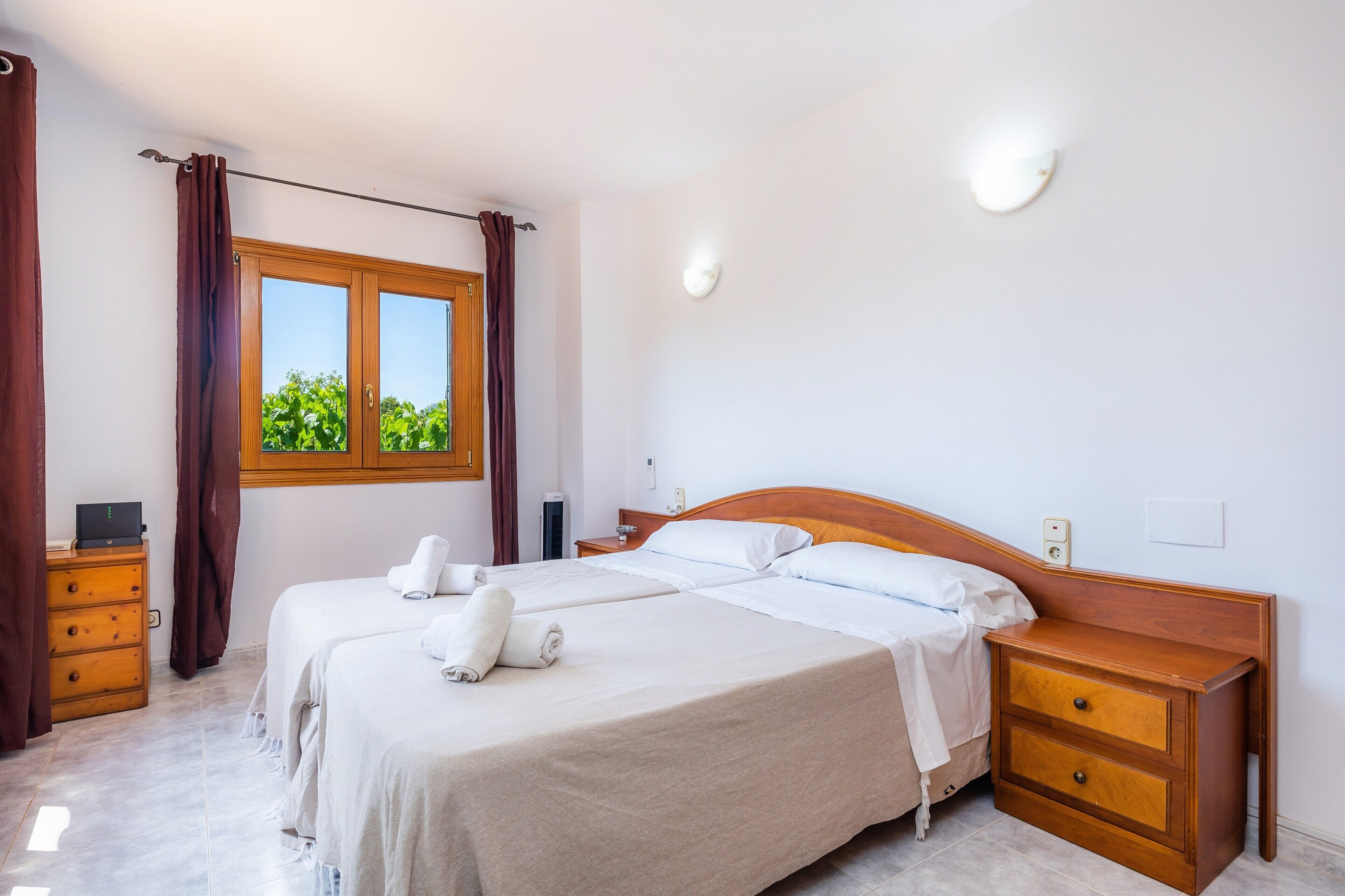 Habitación de hotel con dos camas, una mesita de noche de madera, un aparador, una ventana con cortinas y una lámpara fijada a la pared.