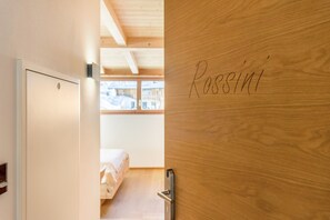 Une porte en bois sur laquelle est gravé le nom « Rosini », une porte blanche avec une poignée, et un aperçu d’une chambre à coucher avec un lit et une fenêtre.