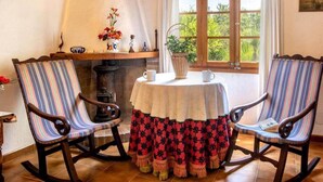 Una habitación con dos mecedoras, una mesa con mantel blanco, un jarrón con flores y una ventana que da a vistas de vegetación.