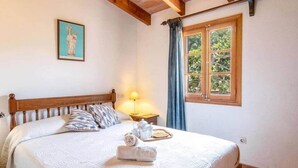 Un dormitorio con una cama de madera, un cuadro en la pared, una ventana con cortinas y una mesita de noche con una lámpara.