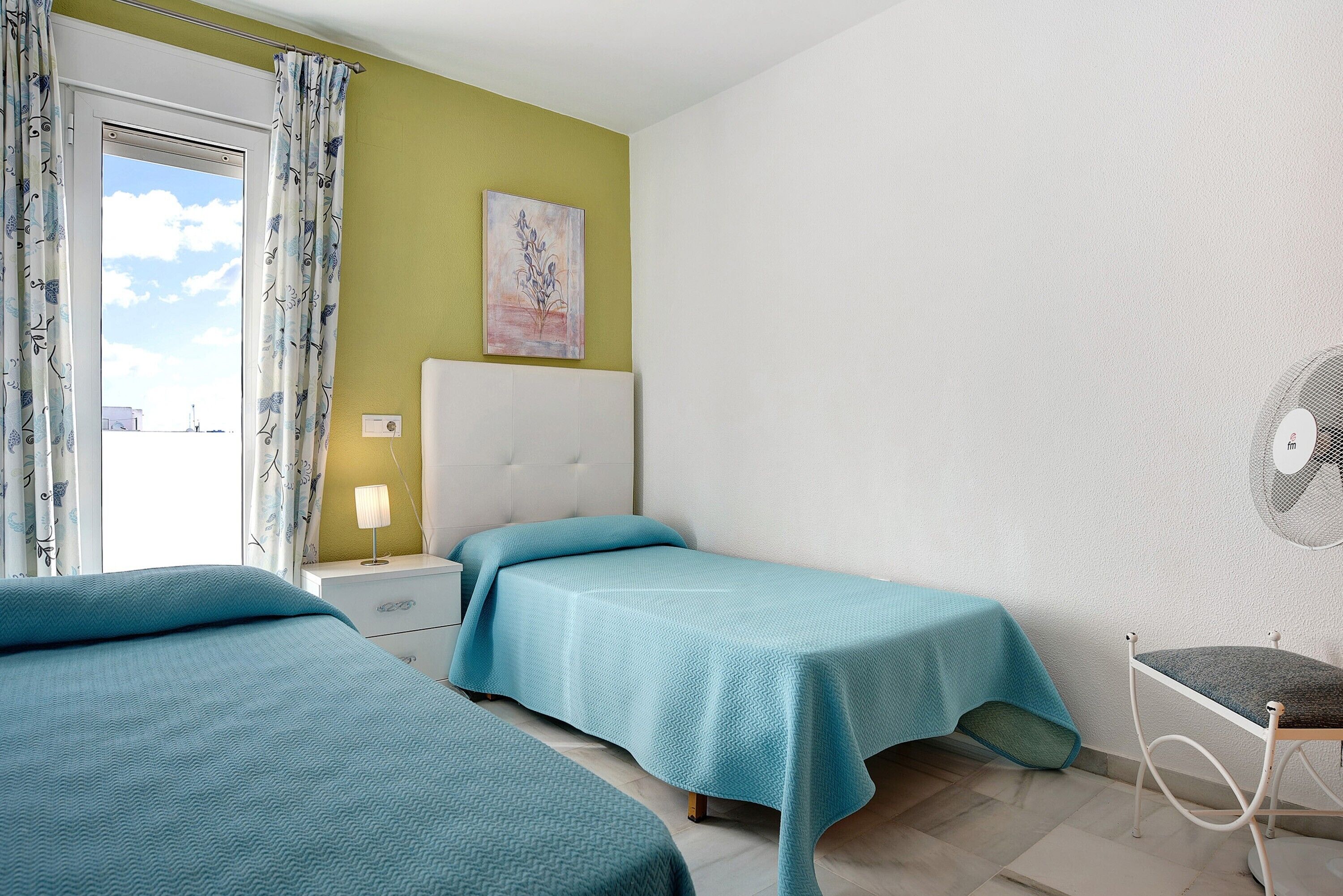 Ein Zimmer mit zwei Betten, einem Nachttisch mit Lampe, einem Sessel, einem Wandventilator und einem Fenster mit Vorhängen.