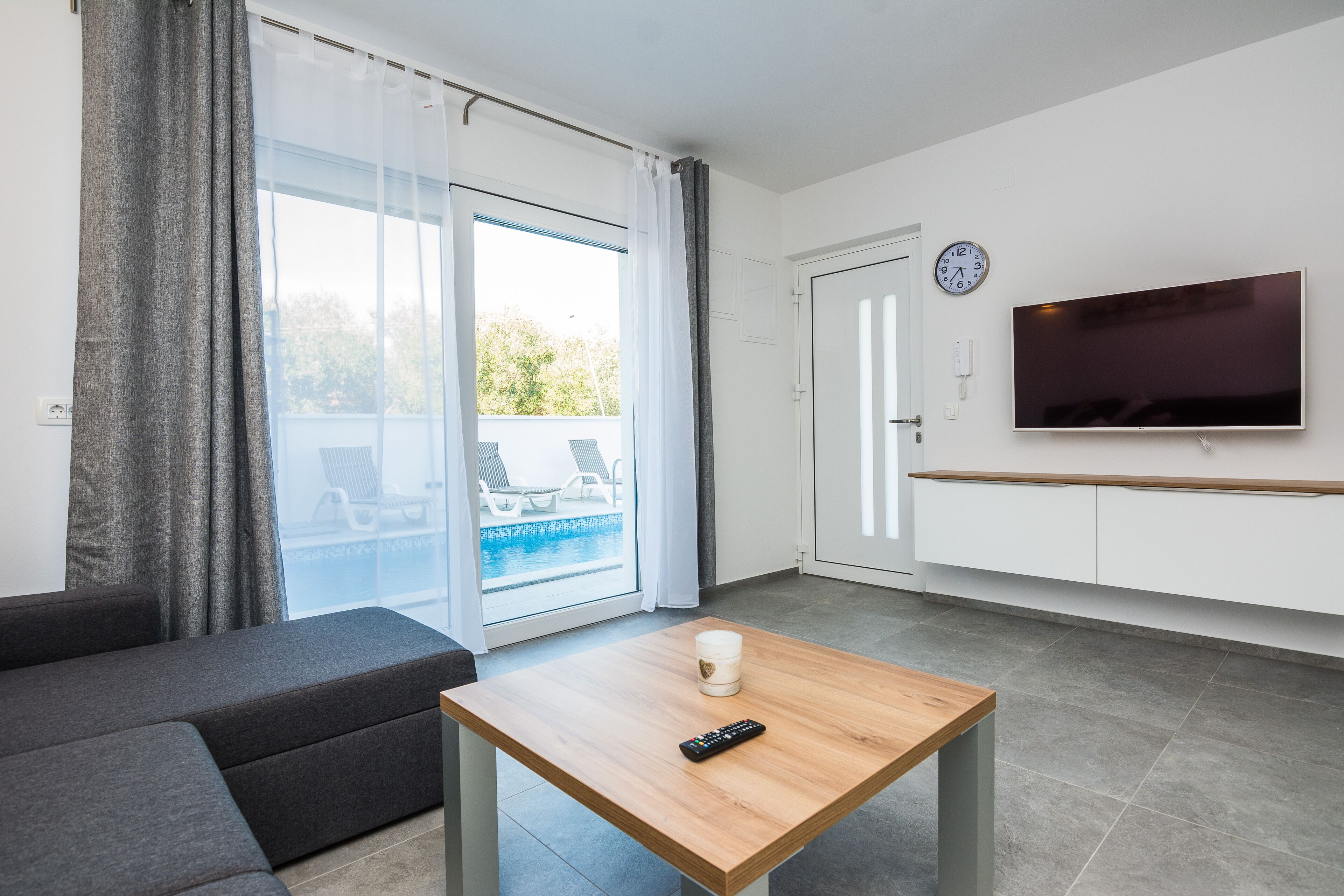 A modern living room with a sofa, a coffee table with a remote, a TV, and a sliding glass door leading to a balcony with a pool.