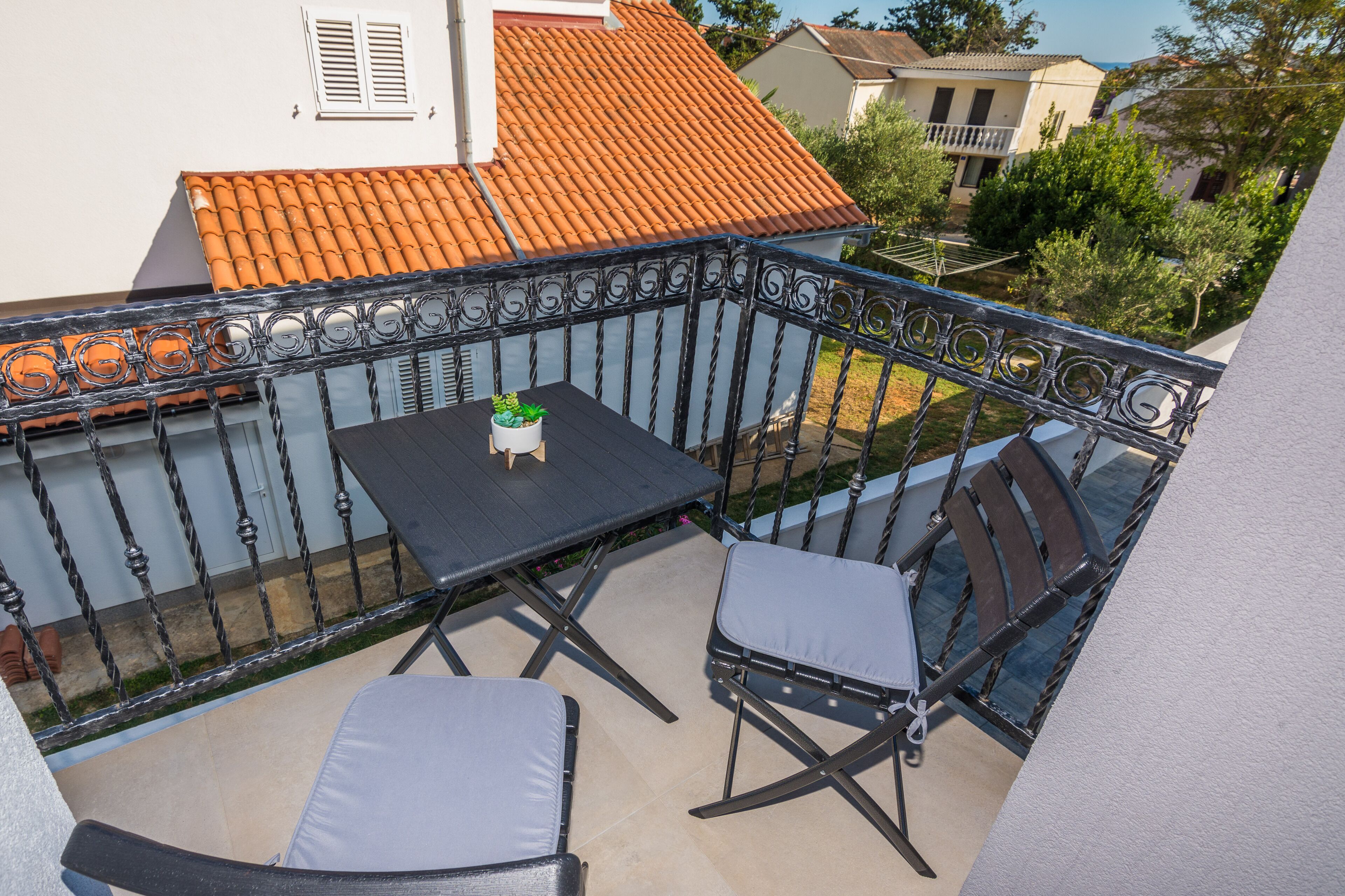 A balcony with a table and two chairs, a potted plant, and a view of neighboring houses.
