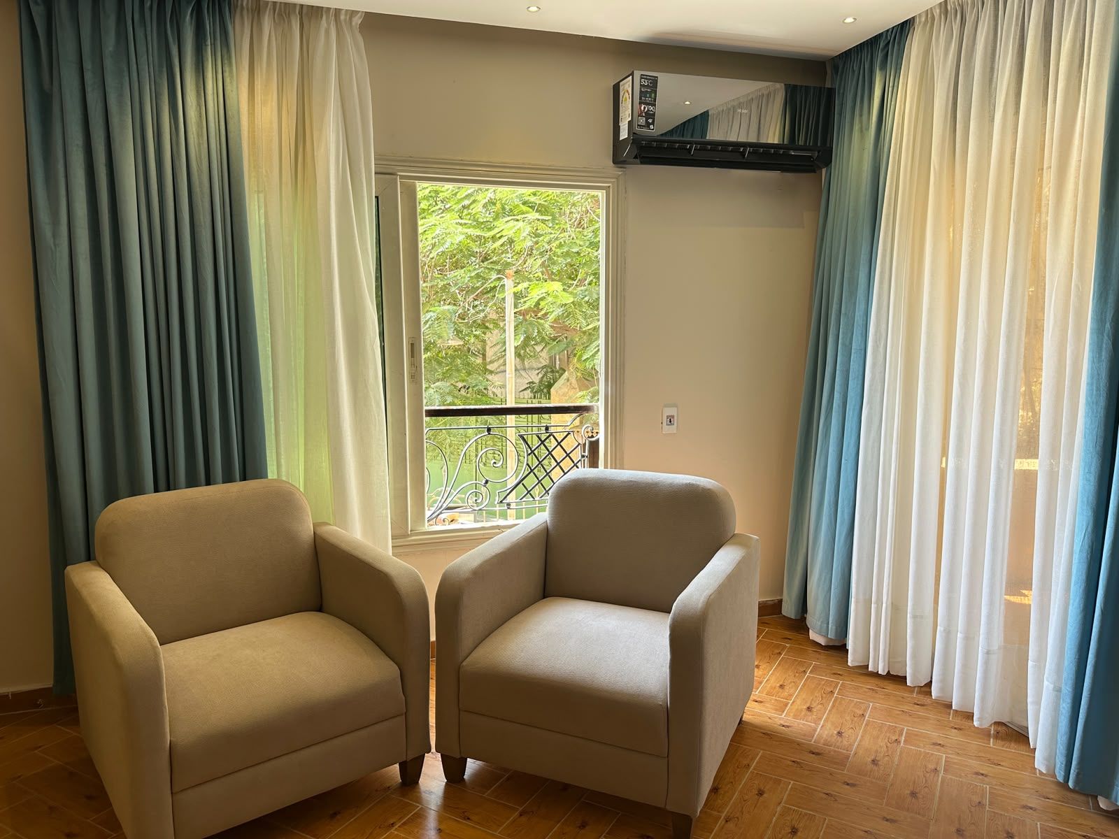 A room with two beige armchairs, a window with a view of greenery, and an air conditioning unit mounted on the wall.