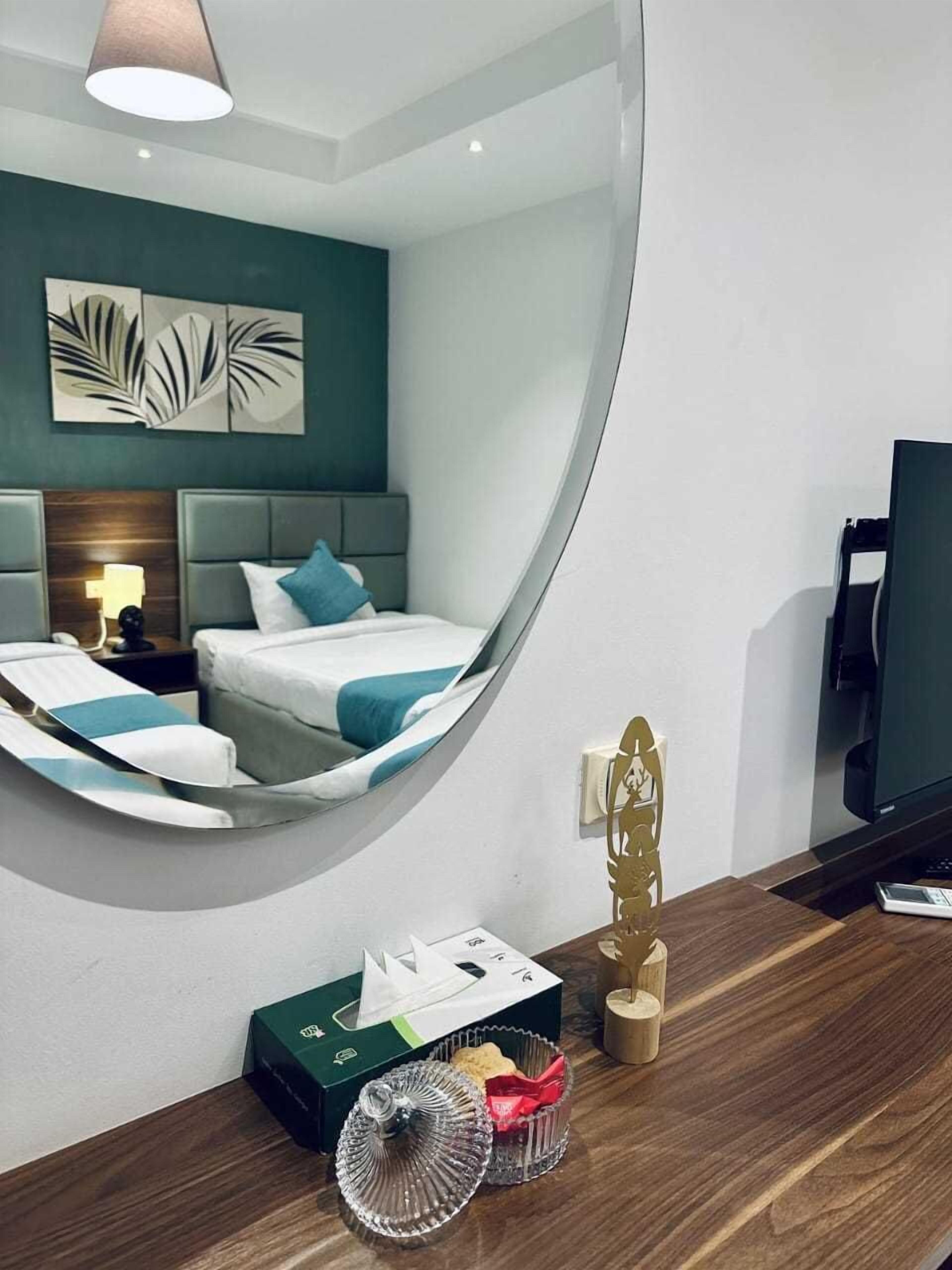 A modern hotel room with a curved mirror reflecting the bed and artwork, a wooden desk with a remote control, and a small table with a tissue box and decorative items.