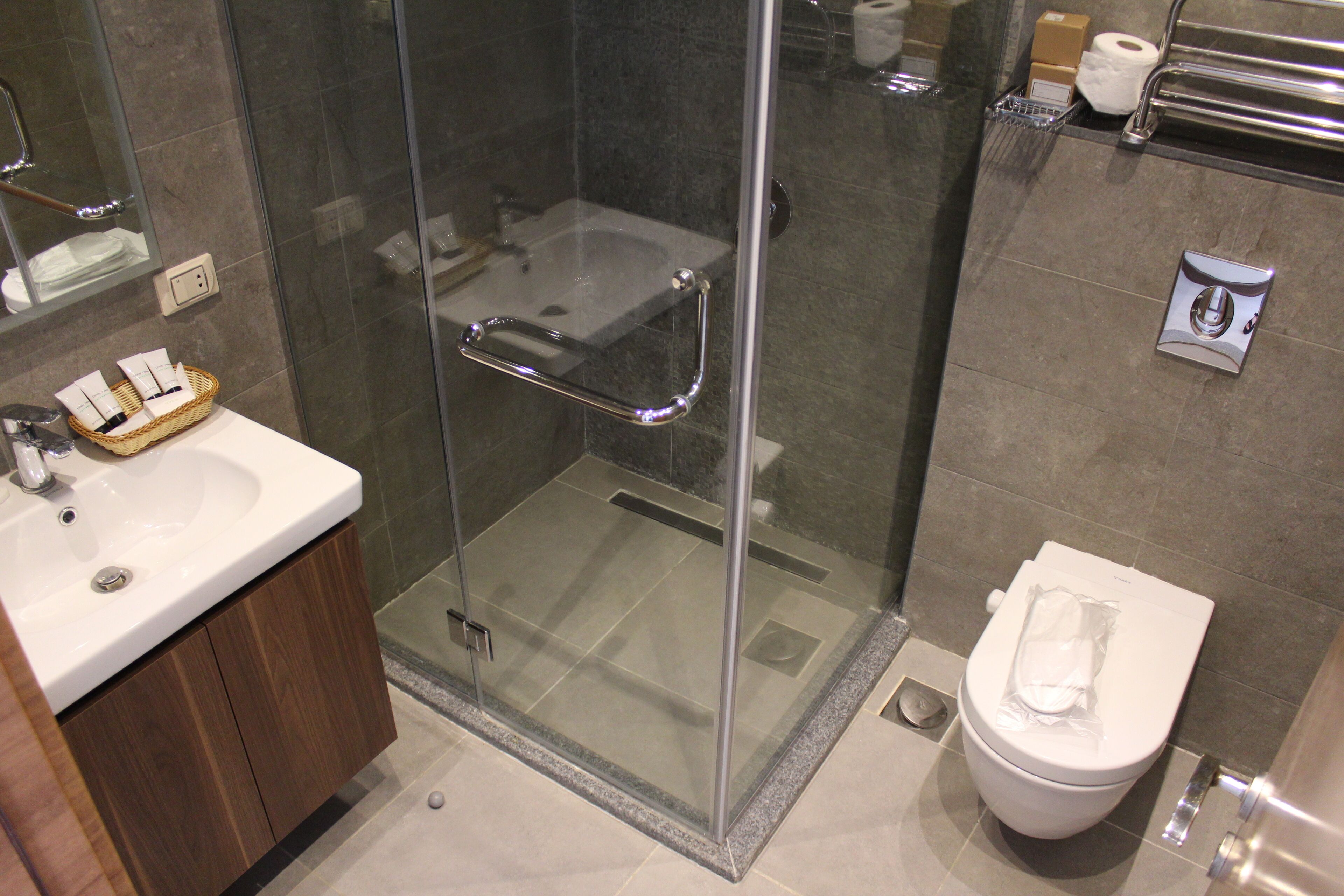 A modern bathroom with a glass-enclosed shower, a white sink with a wooden cabinet, and a toilet with a paper towel dispenser.