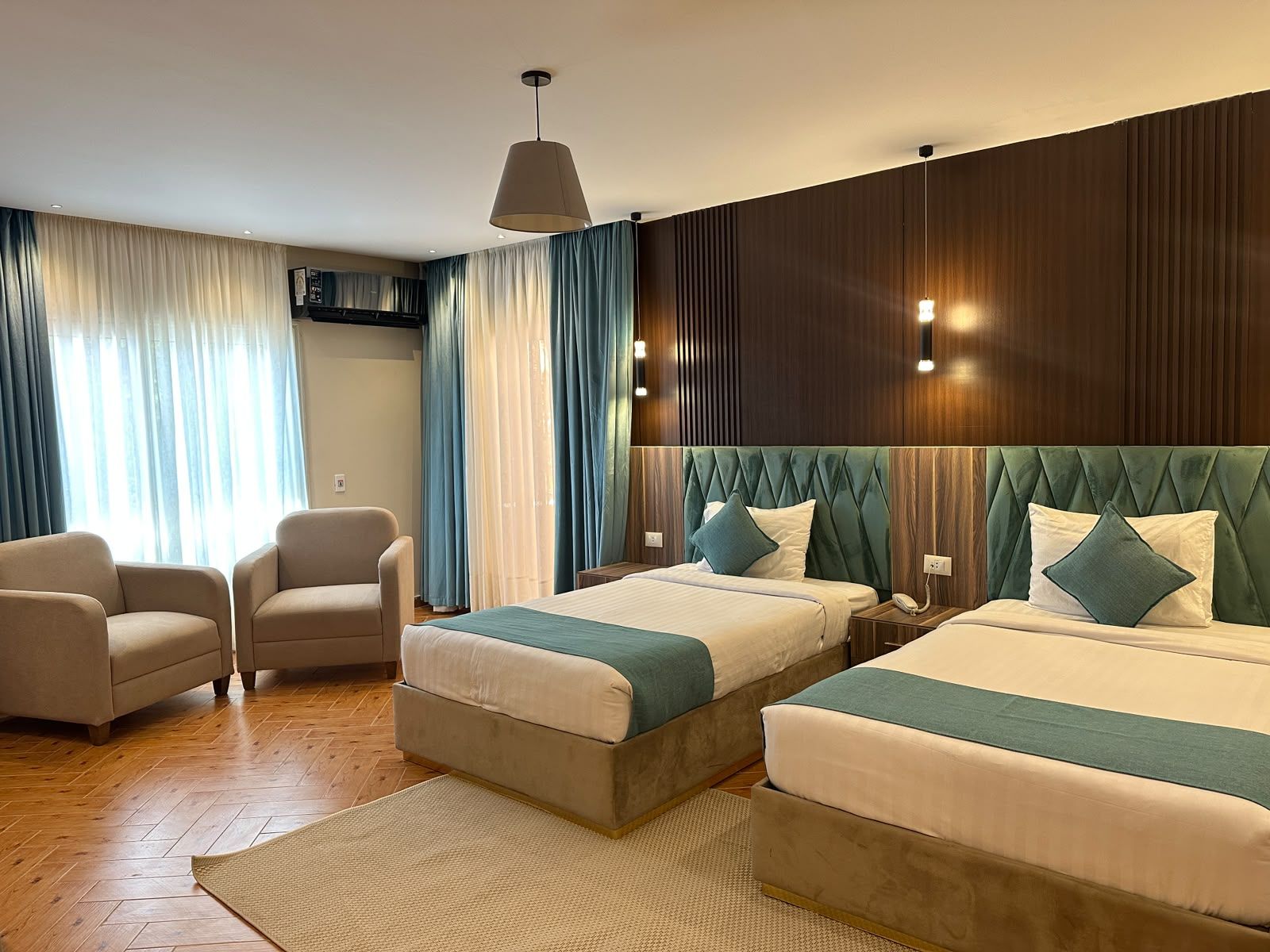 A hotel room with two beds, a sofa, a large window with curtains, and a wooden accent wall.