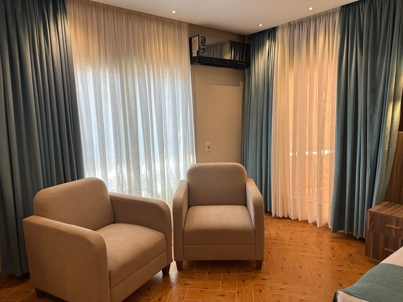 Una stanza con due poltrone beige, pavimento in legno e un letto con coperta verde.