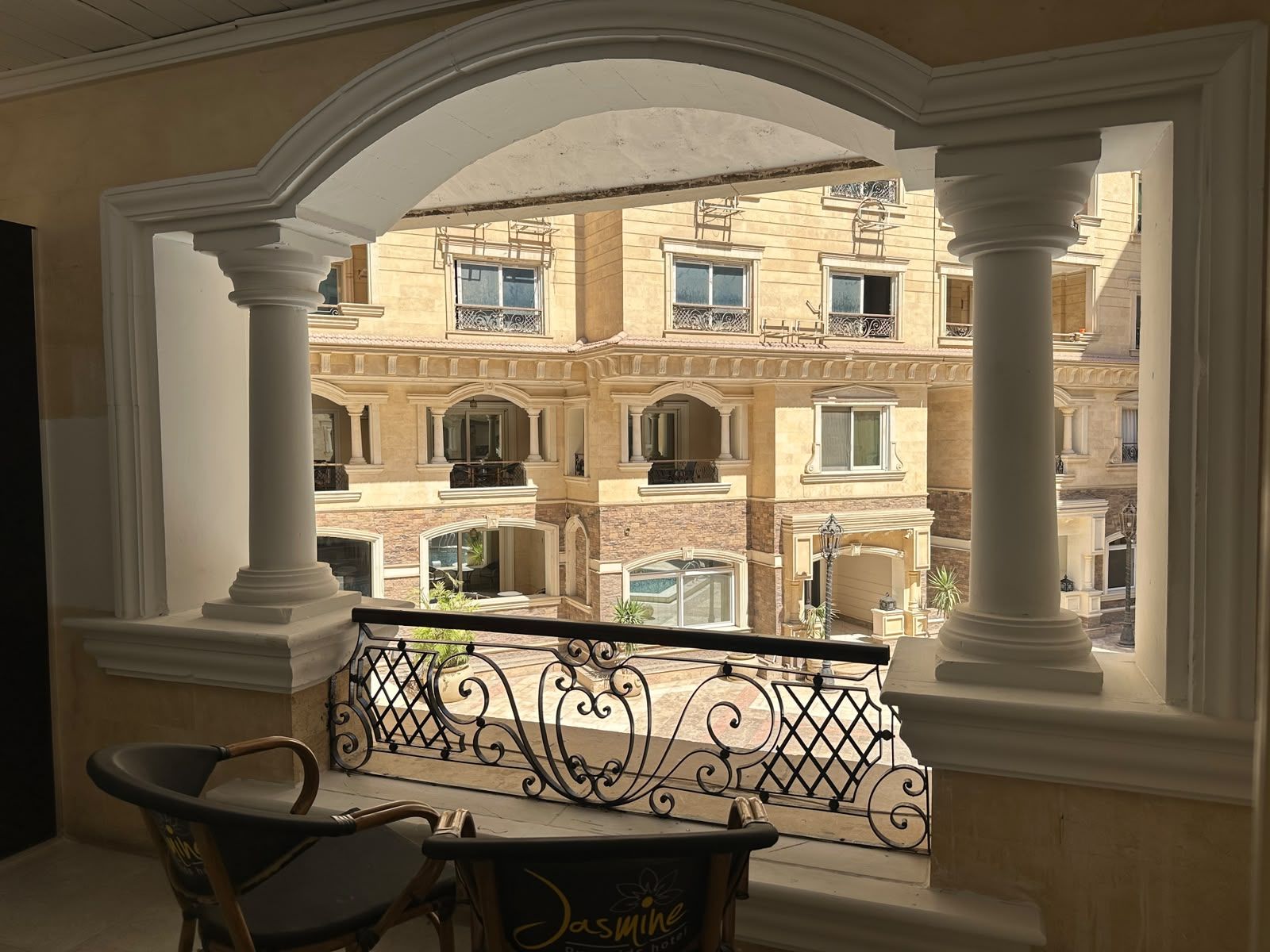 A balcony with ornate metal railings, a chair, and a table, overlooking a building with multiple balconies and windows.