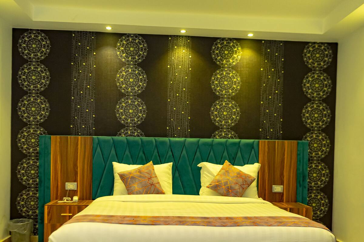 Una cama grande con estructura de madera y cabecera tapizada en verde, una lámpara colgante y una pared con diseños decorativos.