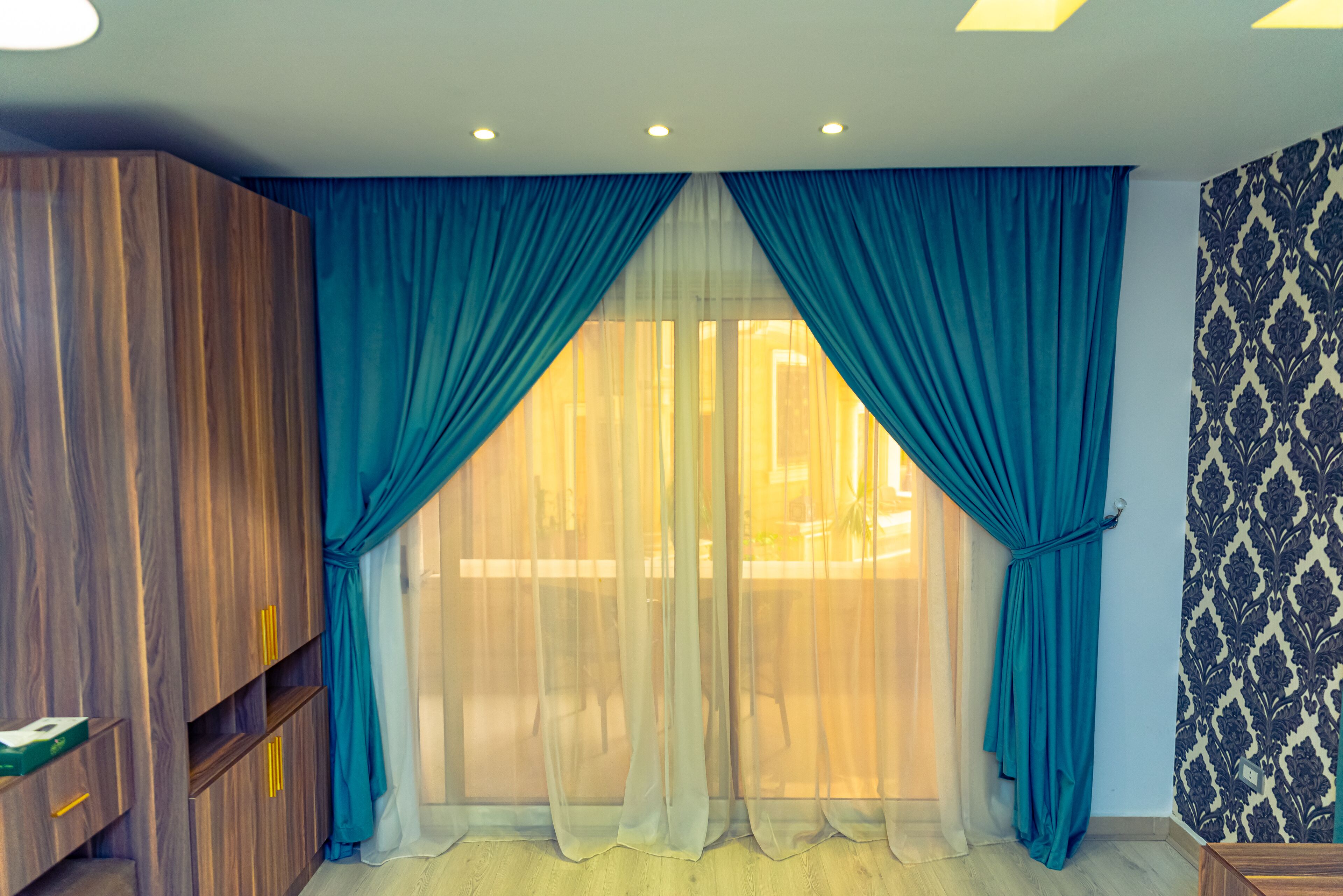 A room with a large window, a wooden wardrobe, and patterned wallpaper.