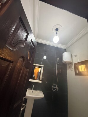 A bathroom with a white sink, dark tiles, a wooden door, a ceiling light, and a shower. - Ideal studio for couples (DAKAR)